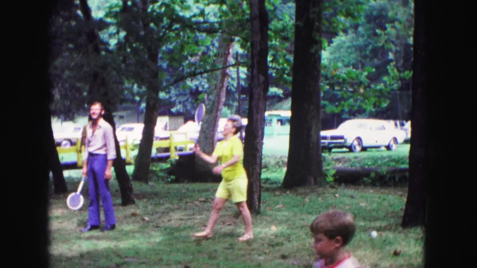 A woman in a yellow outfit swings a racket during a 1967 badminton game in a Galena wooded park while a man watches nearby.