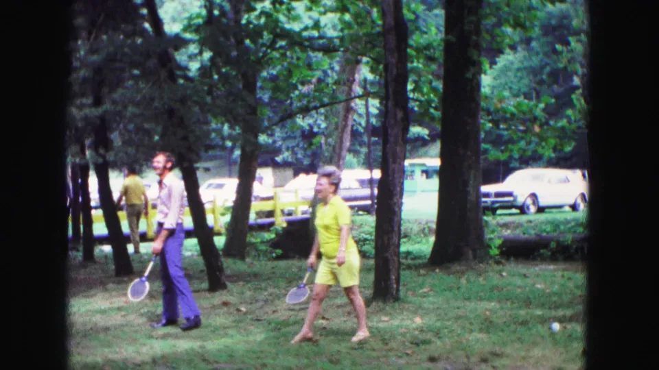 At the end of the 1967 Galena park clip, two badminton players move across the grass with trees, cars, and a yellow fence behind them.