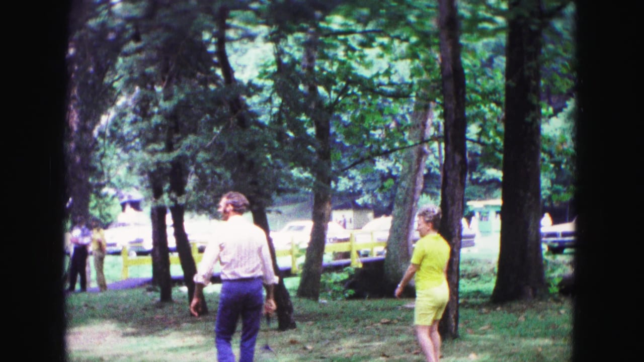 Adults walk through a shaded grassy park with tall trees and benches in the background.