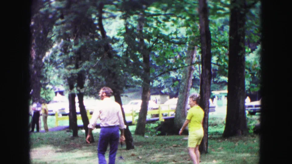 1967 Galena, Illinois 8mm frame showing adults walking through a leafy park under tall trees.