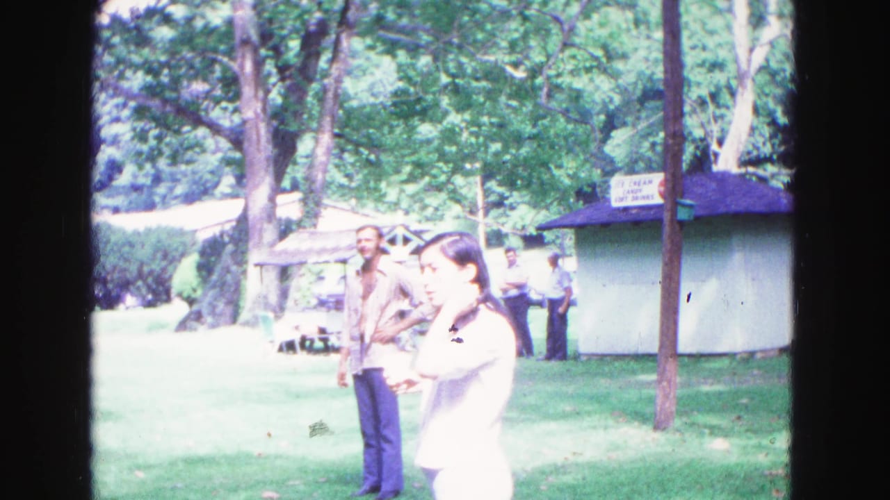 A woman in a white top stands near a man in a patterned shirt by a small park kiosk.