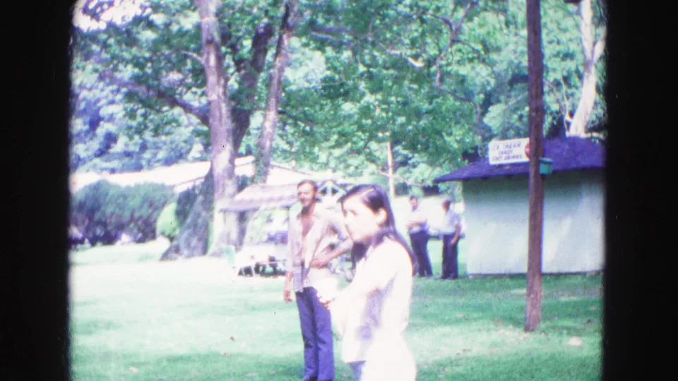 1967 Galena, Illinois 8mm frame of adults in a sunny park beside a kiosk and trees.