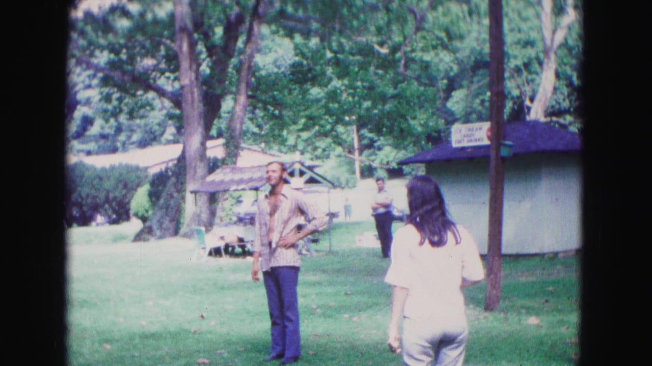 In 1967 Galena, Illinois, adults gather on a park lawn as a man watches a woman near a small concession stand.
