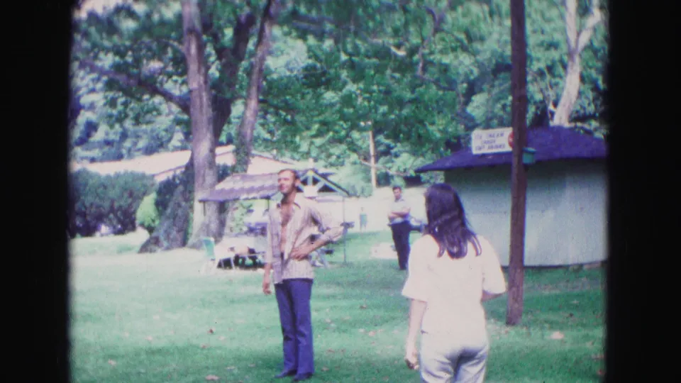 In 1967 Galena, Illinois, adults gather on a park lawn as a man watches a woman near a small concession stand.