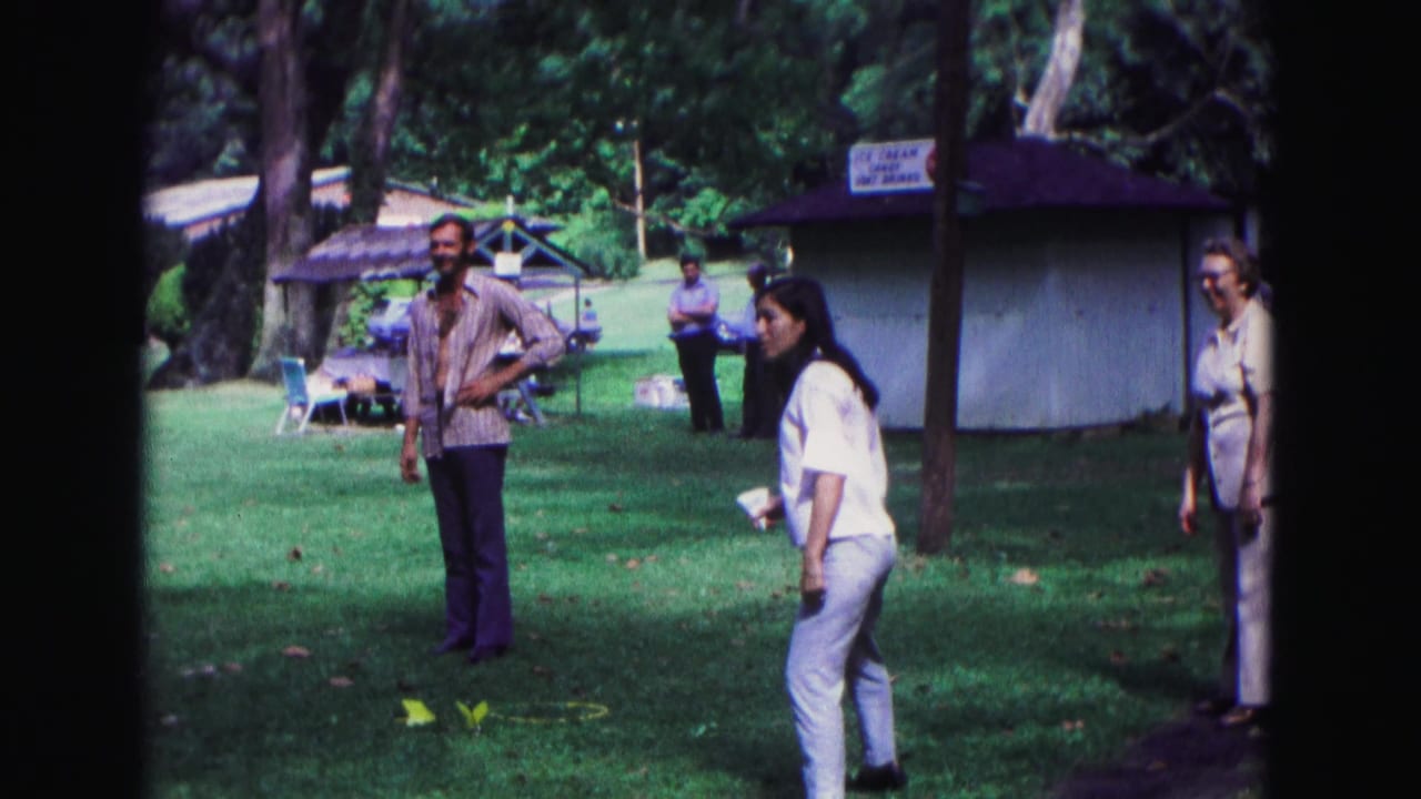 Adults at a 1967 Galena park event watch or take part in a lawn activity near a concession stand and trees.