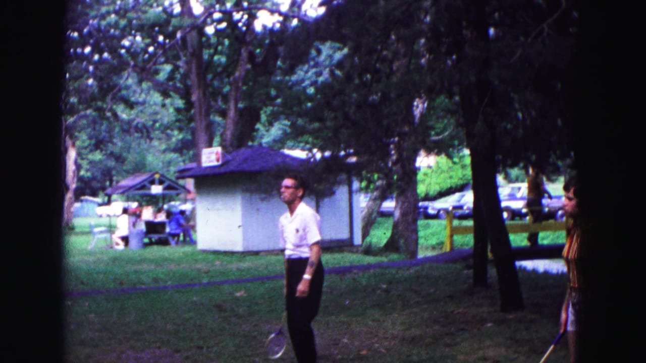 Man with a racket walks on grass near trees and a small park building.