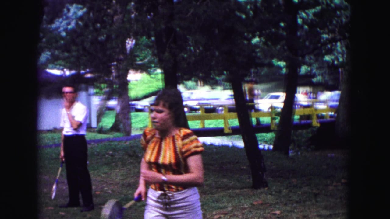 A woman in a striped top and white pants holds a racket in a 1967 Galena park scene, with a man and parked cars behind her.