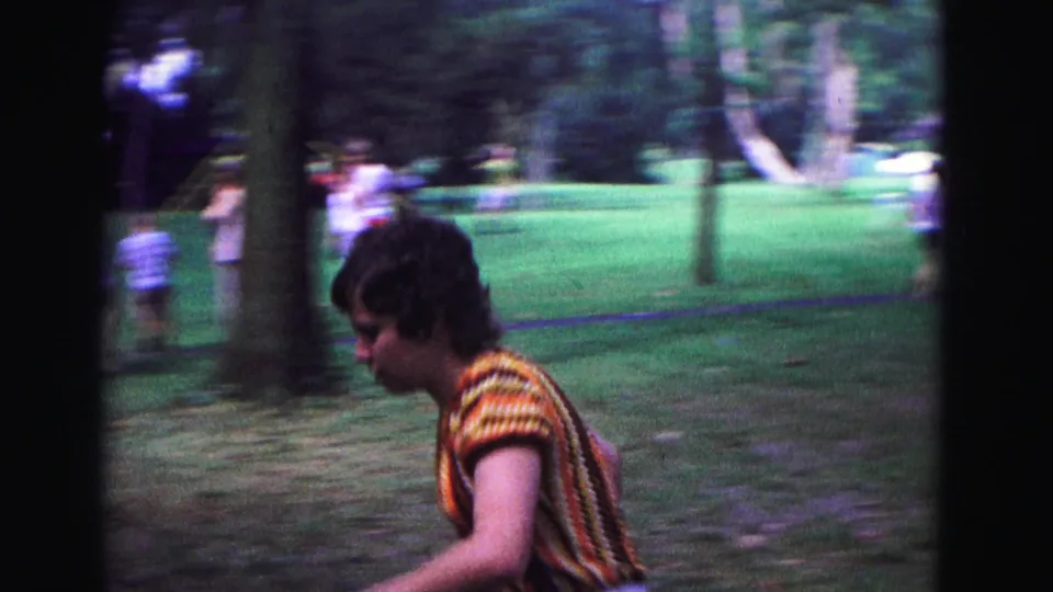 The camera follows a woman in striped 1960s clothing as she moves through the park with a racket in hand.