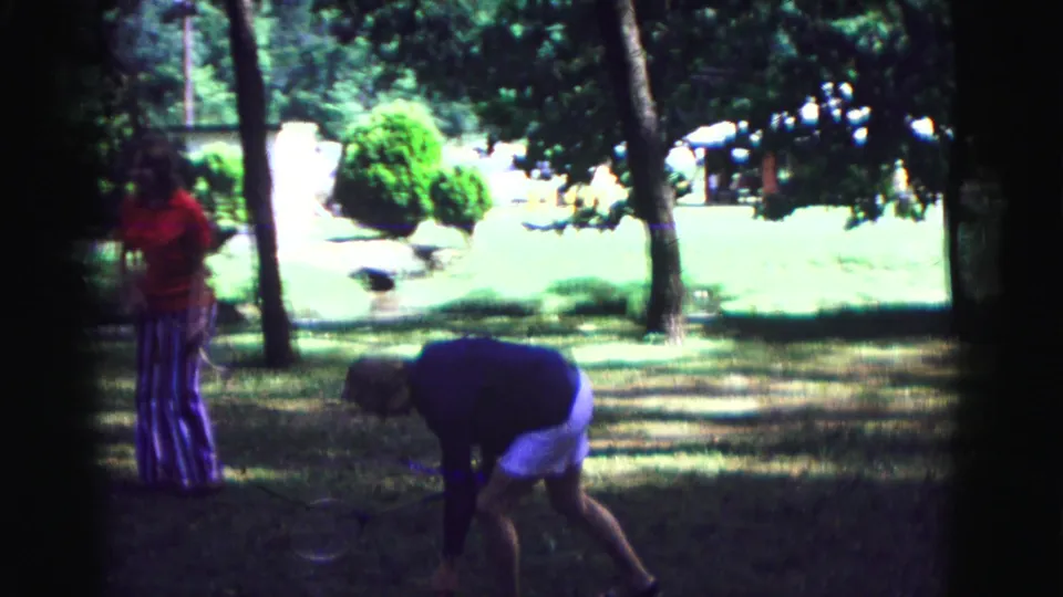 Two women in a Galena park scene stand near badminton equipment, with one bending toward the ground under trees.