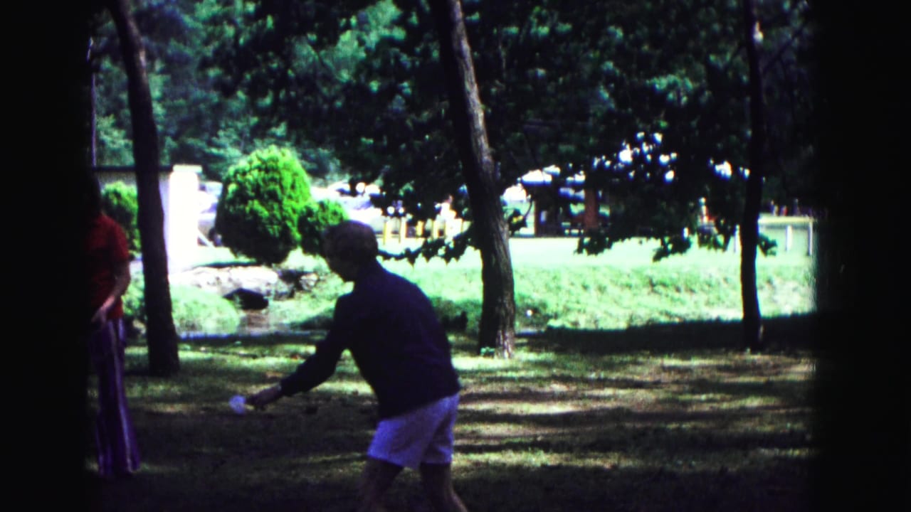 Adults in a shaded park area hold tennis rackets near trees and bright lawn.