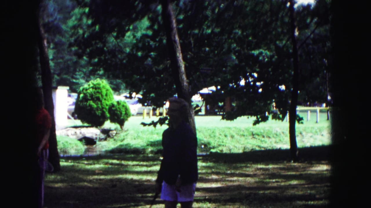 A person walks across a shaded park lawn with trees and bright greenery behind.