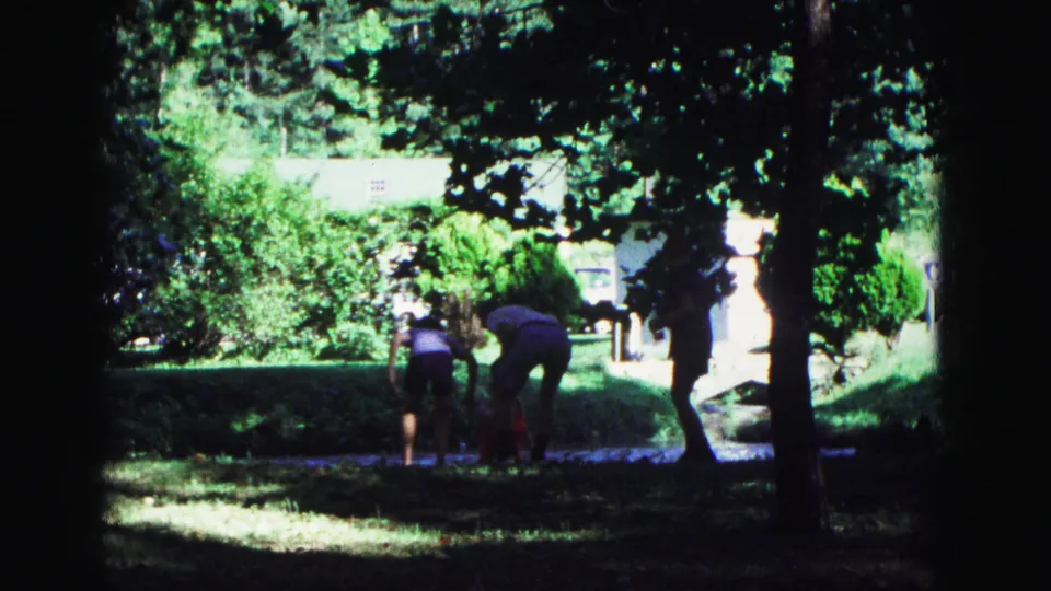 1967 Galena home-movie frame with family members gathering near rackets and items on the grass under trees in a park.