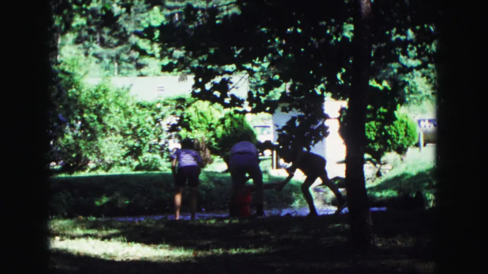1967 Galena park frame showing family members reaching toward the grass near tennis equipment, with trees and shrubs in strong sunlight behind.