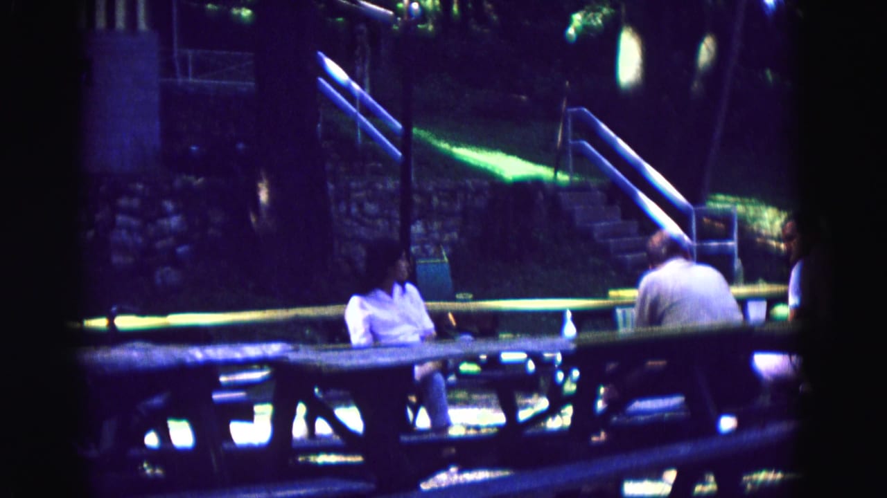 Adults sit at a picnic table near stone steps and blue railings in dim outdoor light.