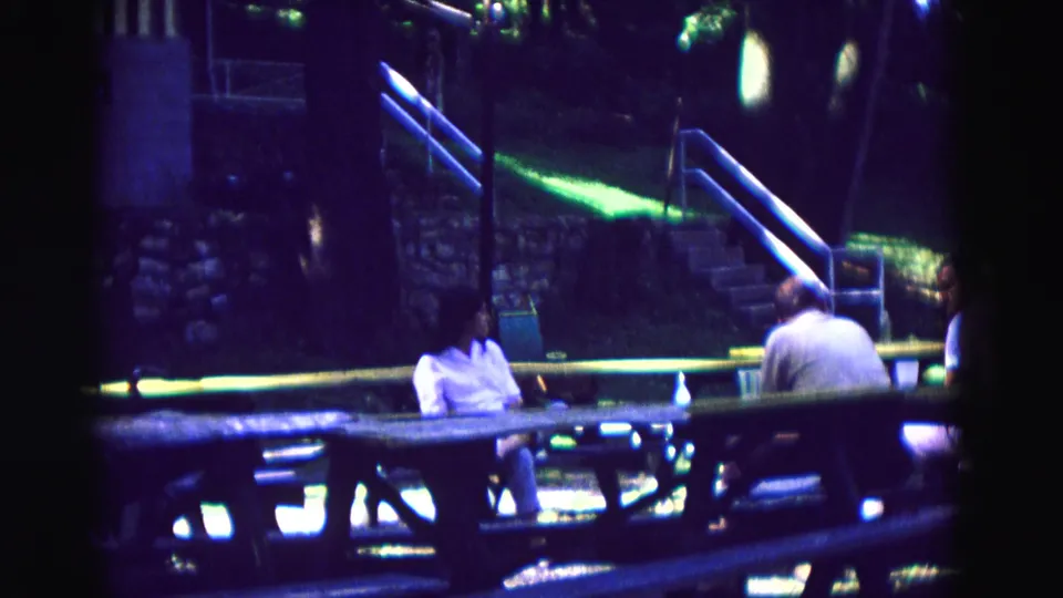 1967 Galena, Illinois home-movie frame with adults seated at a picnic table near stone steps, benches, railings, and trees at night.