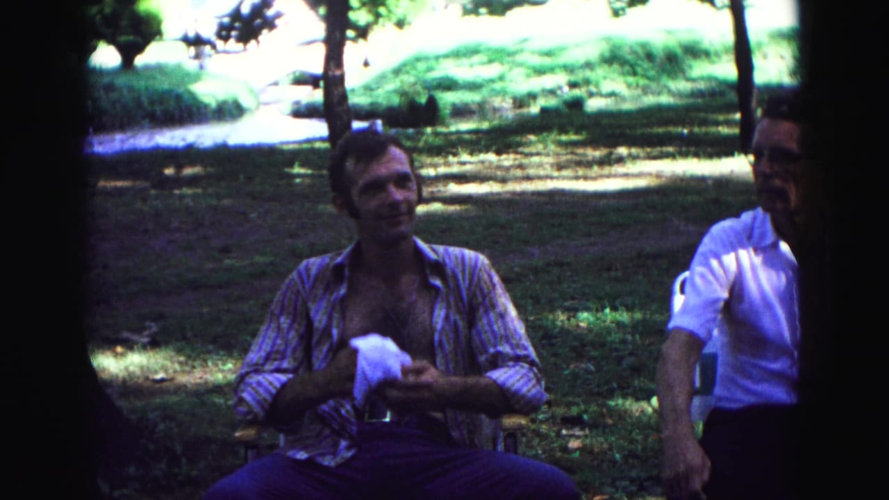 1967 home-movie frame from Galena, Illinois, showing two adult men seated in a shaded park area with trees and grass behind them.