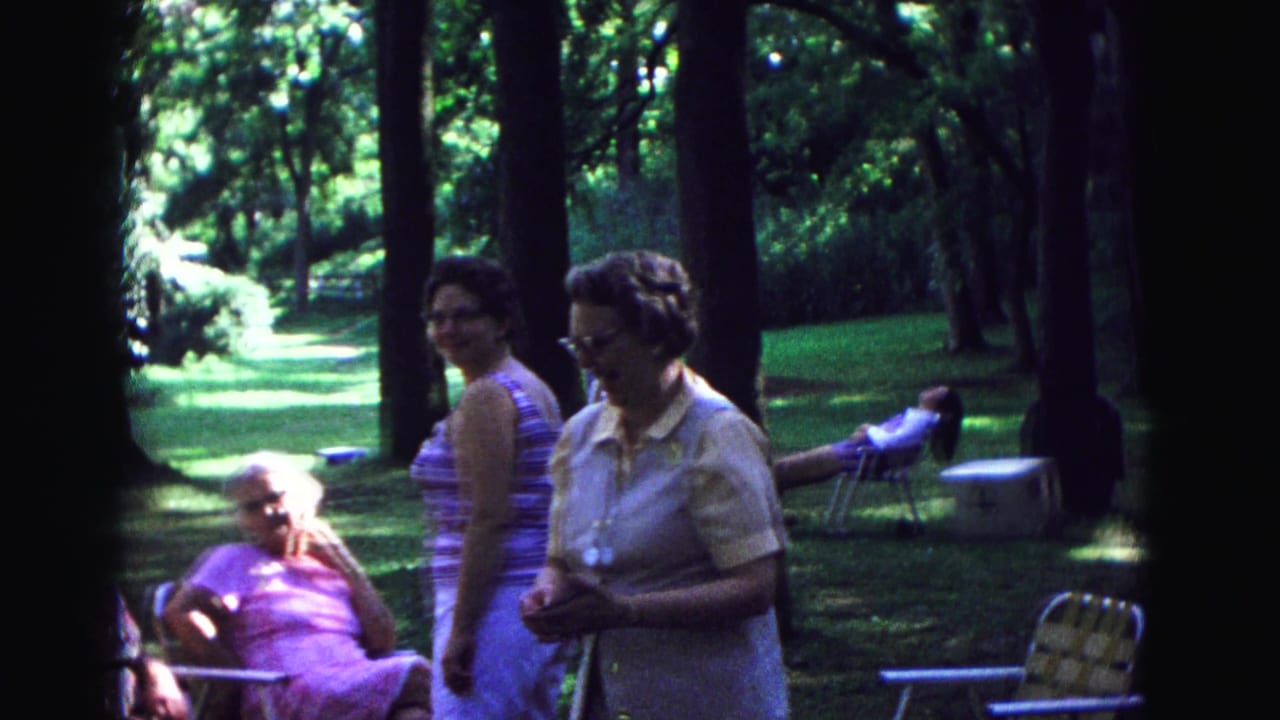 Adults stand and sit among folding chairs in a shaded park.