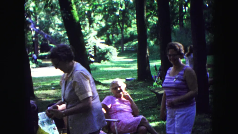 1967 Galena home-movie frame showing women in summer clothing near an older woman seated in a lawn chair under trees.