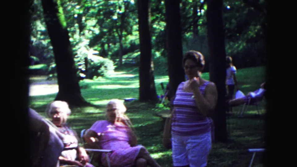 Final 1967 Galena park frame with a woman standing in front of older adults seated in lawn chairs beneath tall trees.