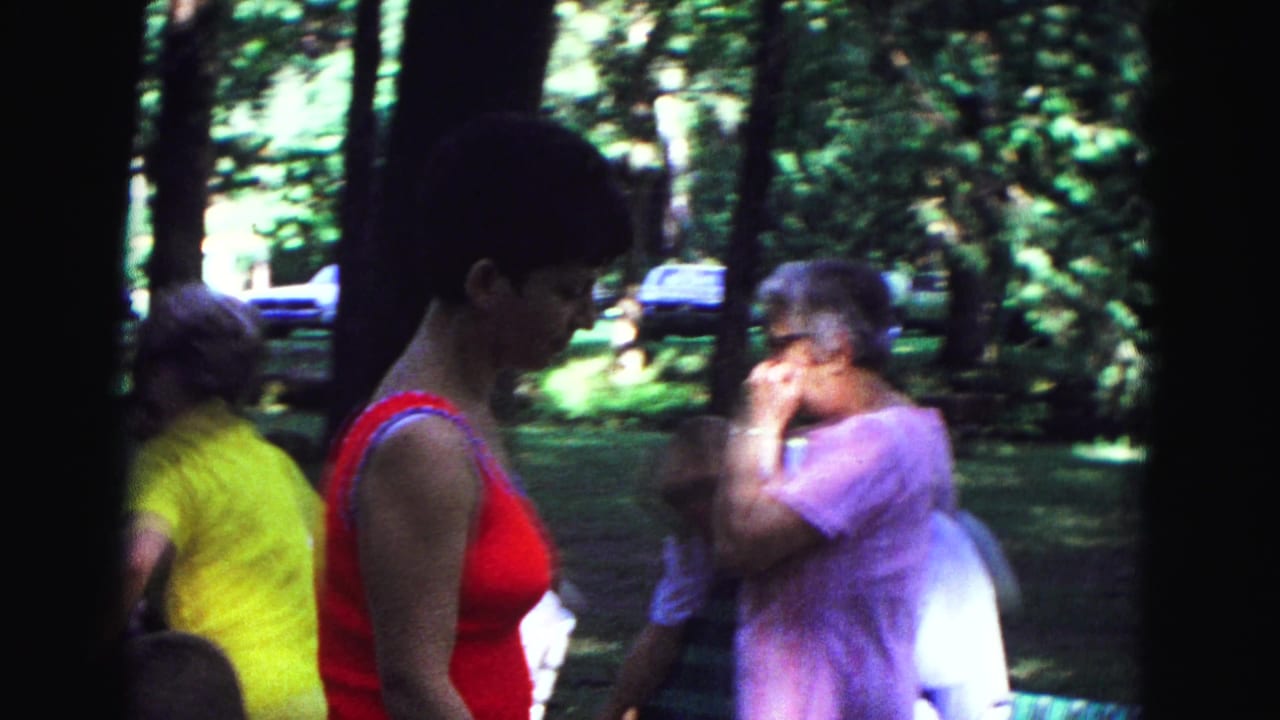 Woman in a red sleeveless top stands near older relatives in a shaded wooded park.