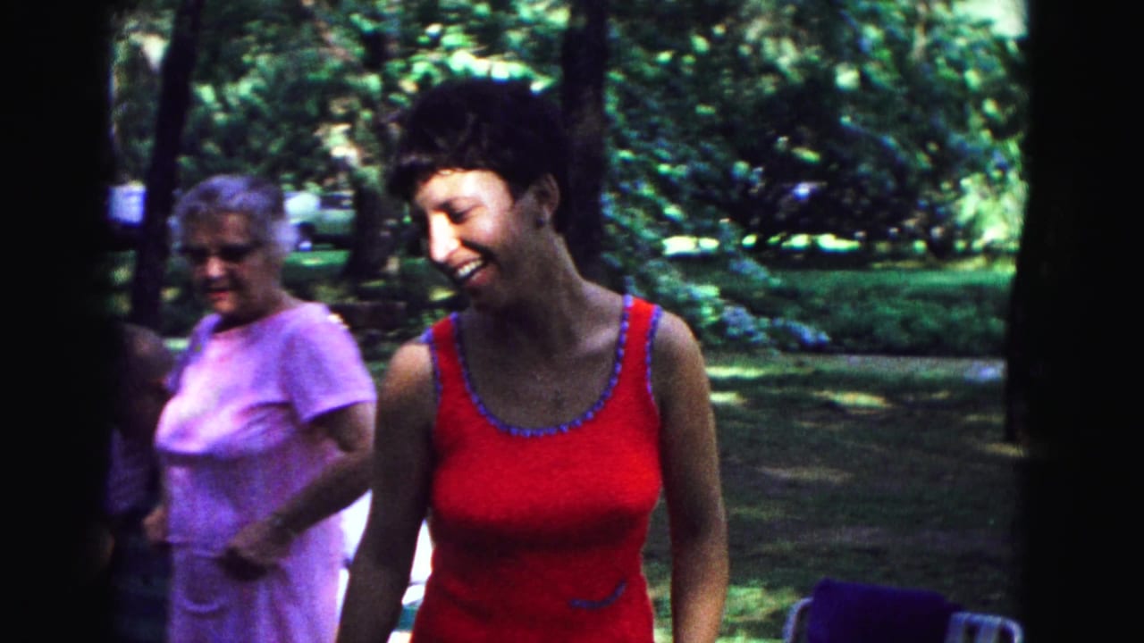 Smiling woman in a red sleeveless top stands near an older woman in lavender under trees.