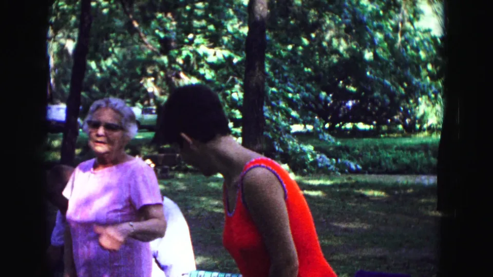1967 Galena home-movie frame showing an older woman in lavender and a woman in red bending near seated relatives in a park.