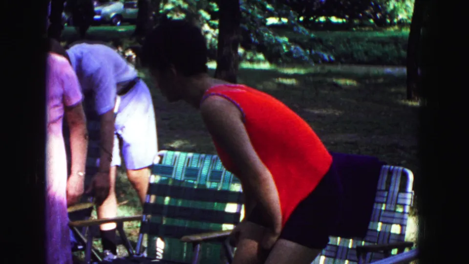 1967 Galena frame showing a woman in red bending near a striped folding lawn chair as relatives gather in a wooded park.