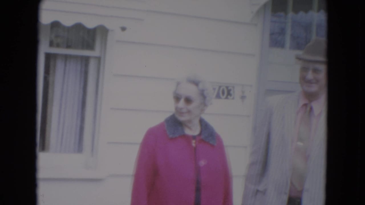 Elderly woman in a pink coat near a man partly cropped at the edge.