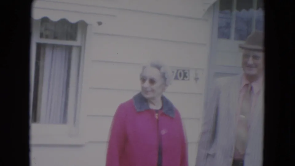 The 1967 Lincoln home-movie frame shows the woman in a pink coat beside the partly visible suited man near a window and awning.