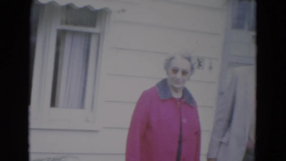 The elderly woman in a bright pink coat smiles near the house window, with the suited man only partly visible at the right edge.