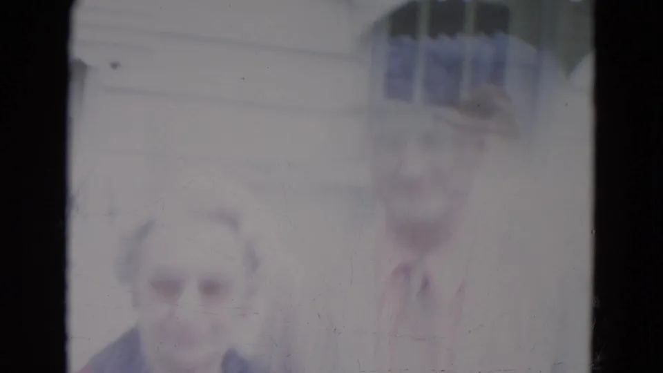 A soft, pale 8mm frame shows the elderly couple close to the camera, with the man in a fedora and the woman in glasses.