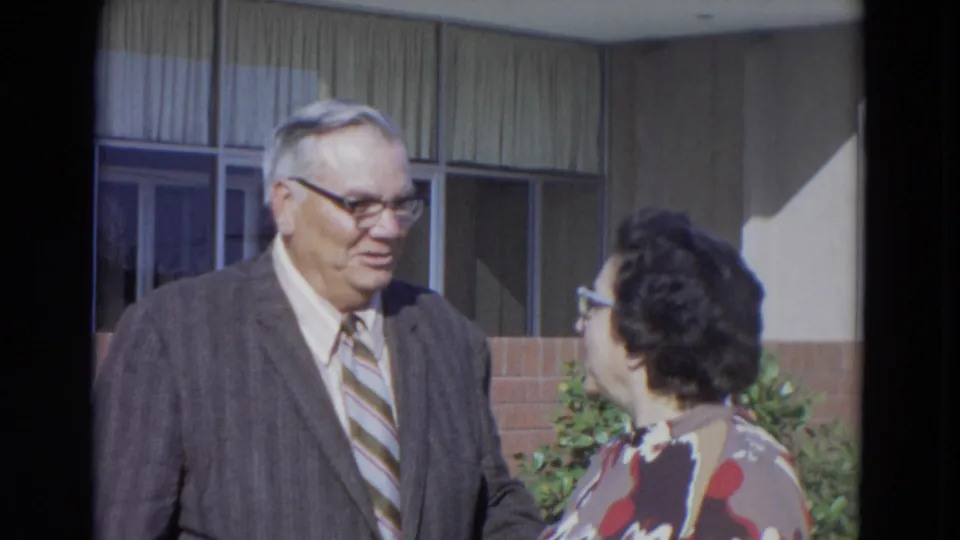 The man and woman turn toward each other during the 1967 home-movie shot, with brickwork, windows, and shrubs visible behind them.