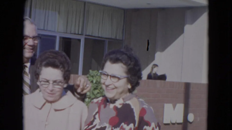 Two women in 1960s clothing smile in front of a brick-and-window facade, with a man partly visible behind them.