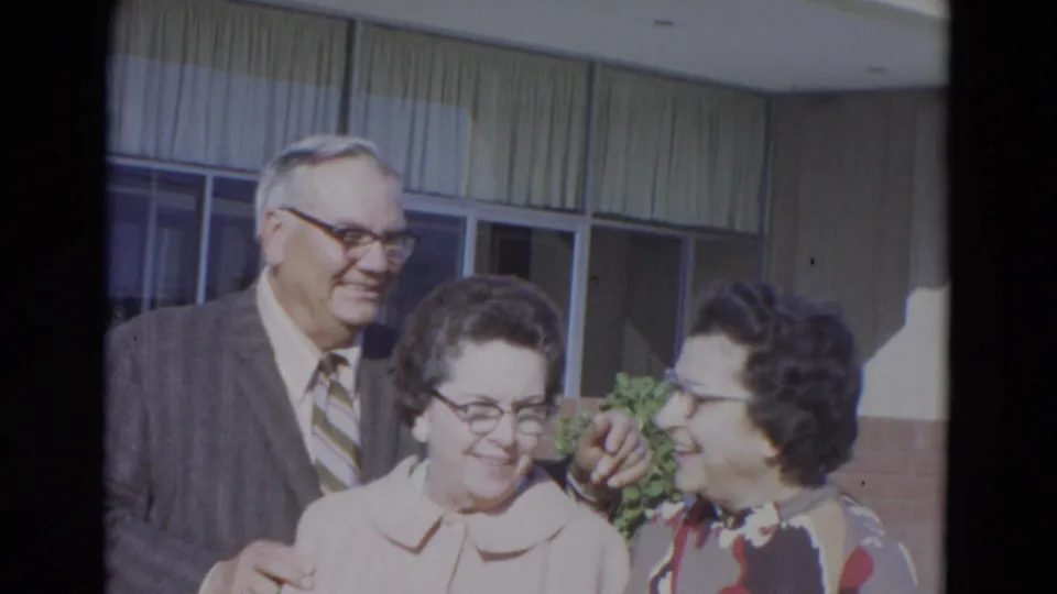 A suited man stands behind two women as one turns toward the other in a candid 1967 Lincoln, Nebraska home-movie moment.