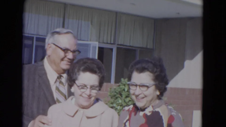 Three adults smile together in a 1967 Lincoln, Nebraska 8mm frame, with brickwork, windows and curtains behind them.