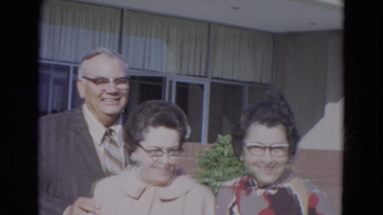 Three older adults wearing glasses pose outdoors near large windows.