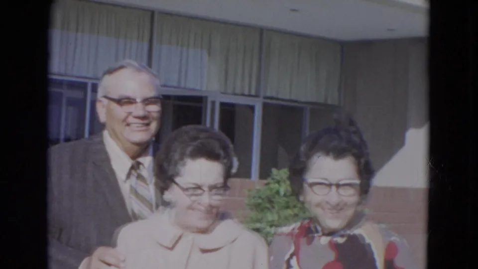 In metadata-supported 1967 Lincoln, Nebraska, three older adults wearing glasses pose in bright sun near a building with large windows.