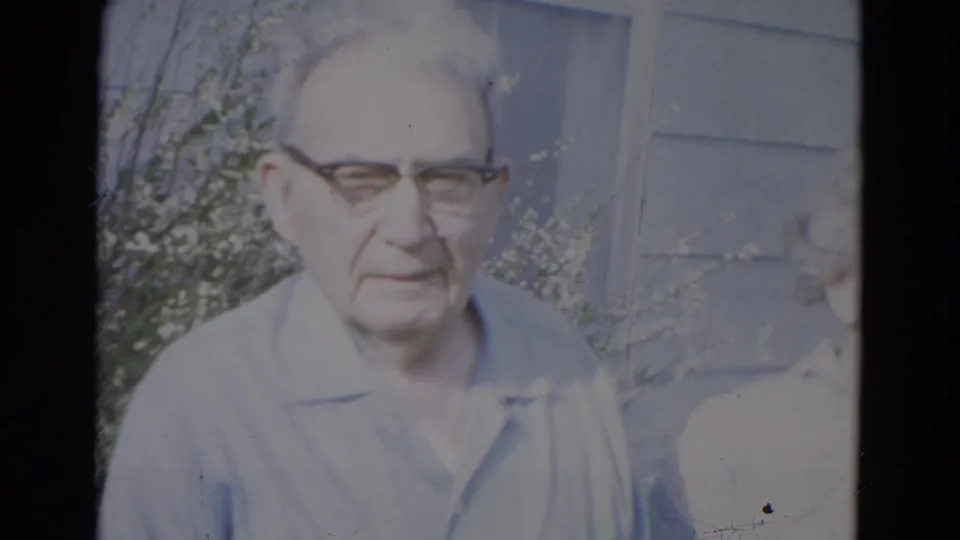 An older man wearing glasses and a light collared shirt stands outdoors near a wall and greenery in the Lincoln, Nebraska home-movie footage.