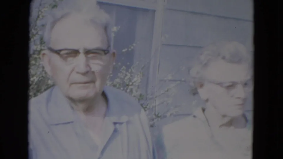 An older man and woman wearing glasses stand close to the camera near an exterior wall and sparse greenery in this 1967 Lincoln, Nebraska clip.