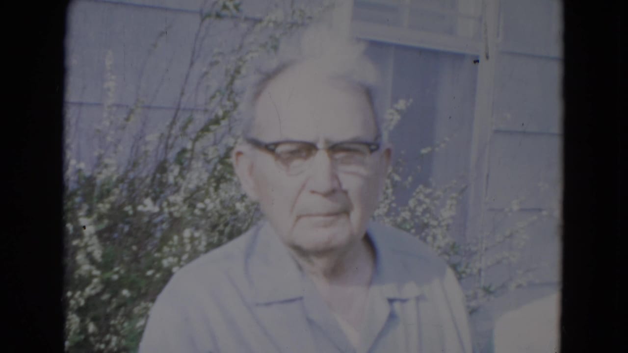 Elderly man in glasses standing outside near gray house siding and shrubs.