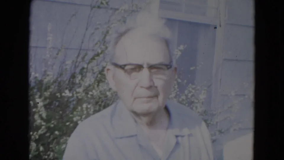 Elderly man in glasses and a light blue collared shirt stands outside a Lincoln, Nebraska home in 1967 8mm footage.