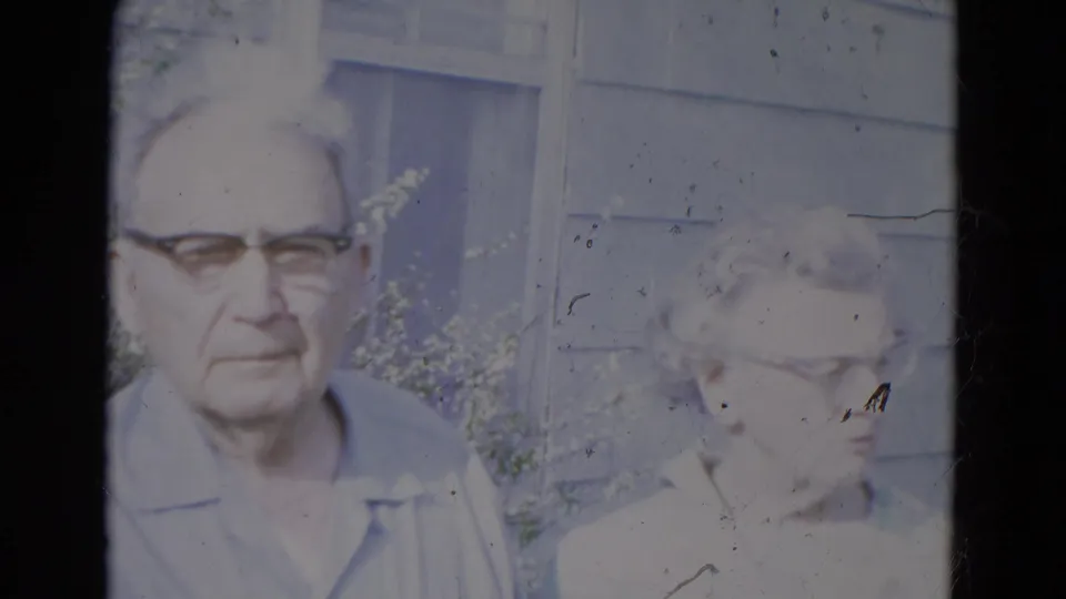 Elderly couple stands outside a gray-sided home in Lincoln, Nebraska, with the woman partly overexposed at frame edge.