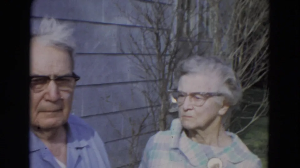 Elderly couple in glasses stands together outside a Lincoln, Nebraska home, framed by gray siding, bare branches, and lawn.