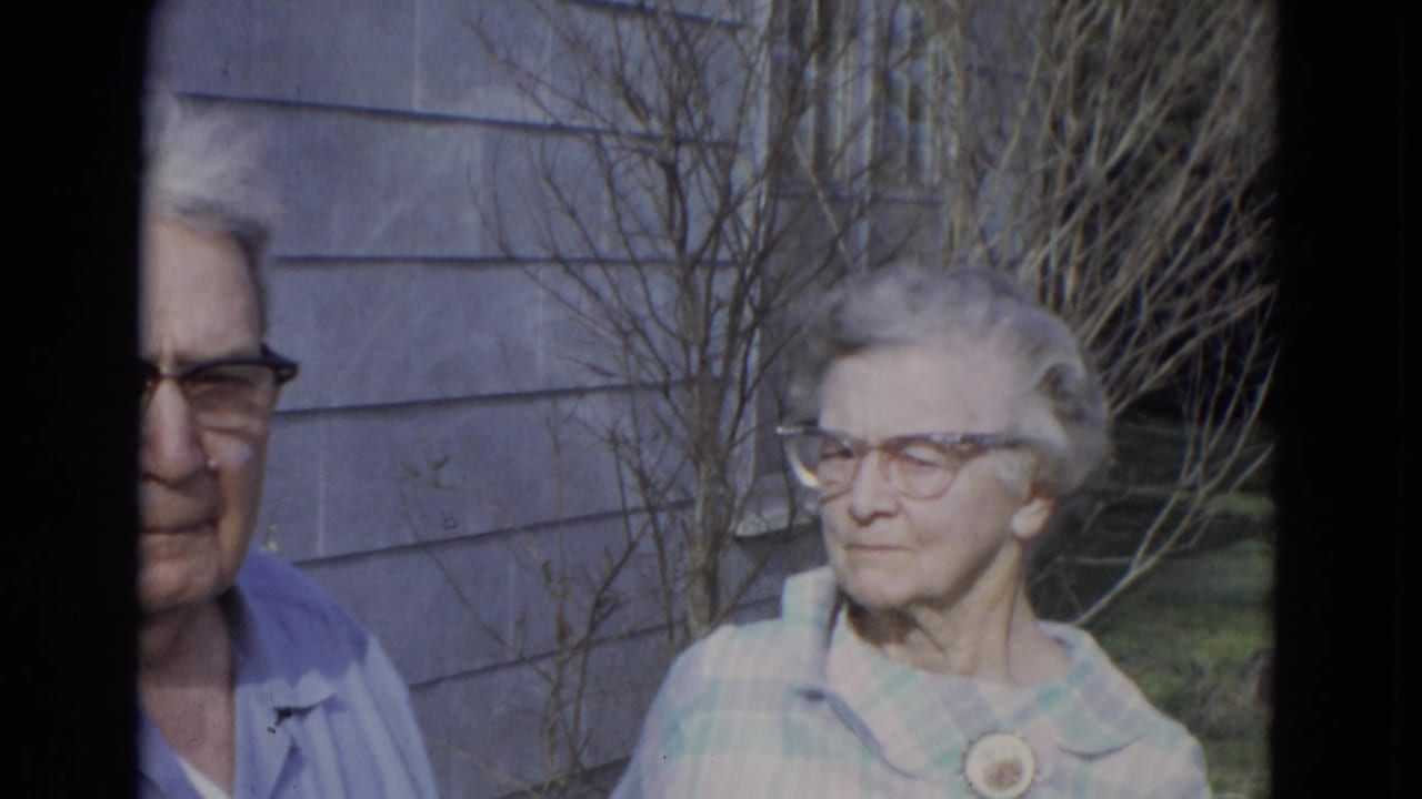 Elderly man and woman wearing glasses stand outside gray house siding.