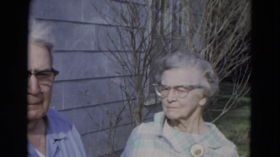 1967 Lincoln, Nebraska home-movie frame of an elderly couple in glasses standing near gray house siding and bare shrubs.