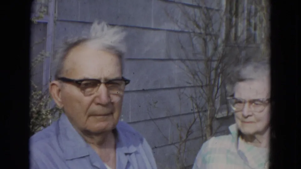 1967 Lincoln, Nebraska frame with an elderly man in a blue shirt centered outside a house and an elderly woman partly visible at right.