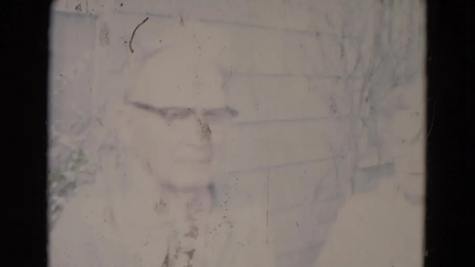 Overexposed 8mm frame from the 1967 Lincoln clip with faint outlines of the elderly couple, house siding, scratches, and dust.