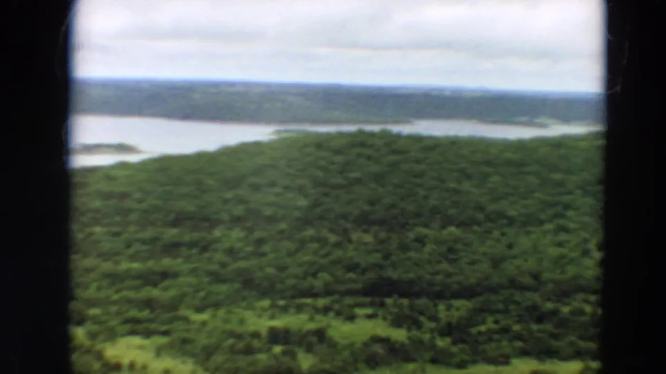 1967 home-movie frame of rolling Minnesota woods overlooking a calm river or lake inlet on a cloudy day.