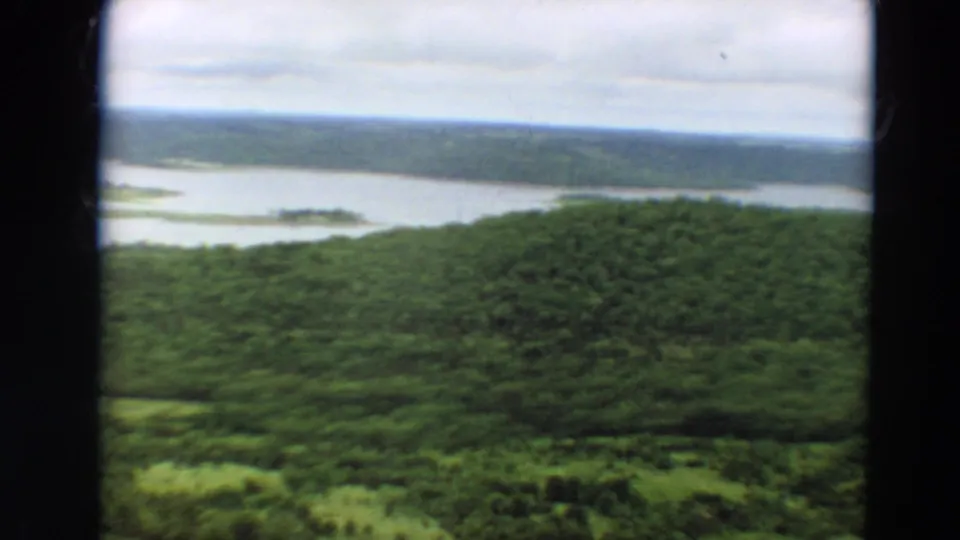 Vintage 8mm Minnesota landscape frame with wooded hills, green foreground, and a calm distant waterway.