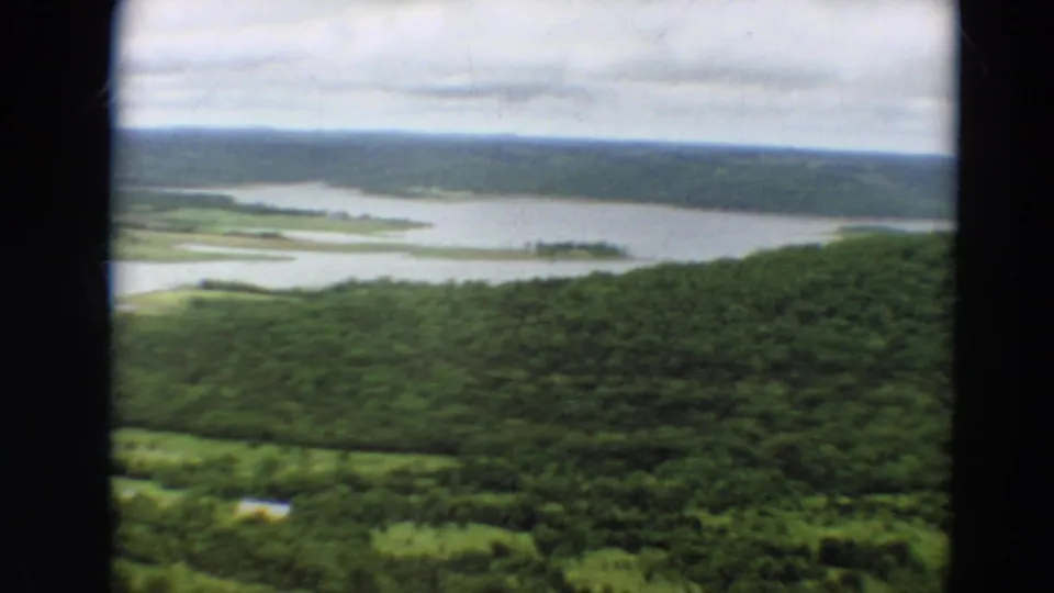 1967 Minnesota home-movie frame with forested slopes, pale water channels, and shoreline clearings seen from an aircraft.